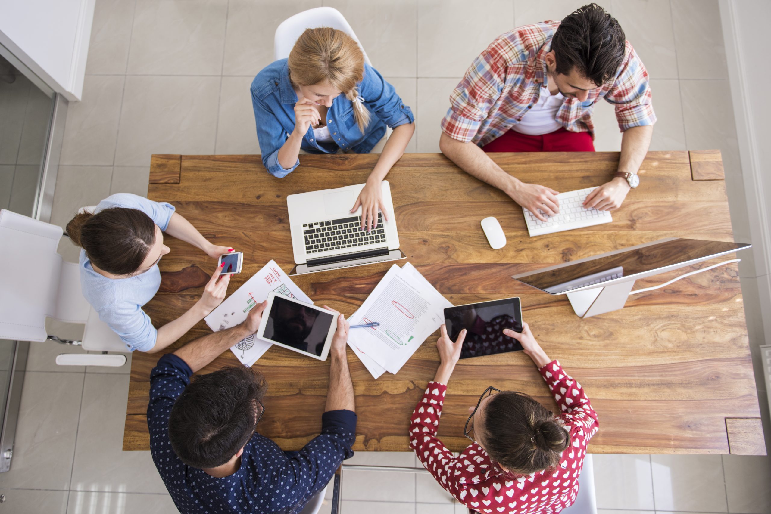 Équipe en réunion autour d’une grande table en bois, utilisant ordinateurs, tablettes et documents de travail.