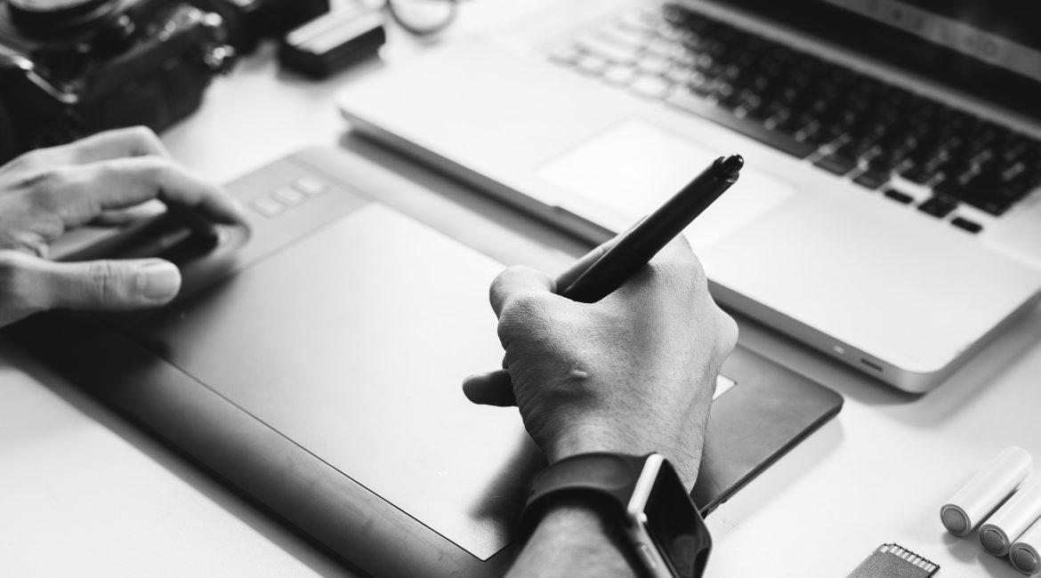 Designer utilisant une tablette graphique et un stylet devant un ordinateur portable, avec du matériel photo sur le bureau.