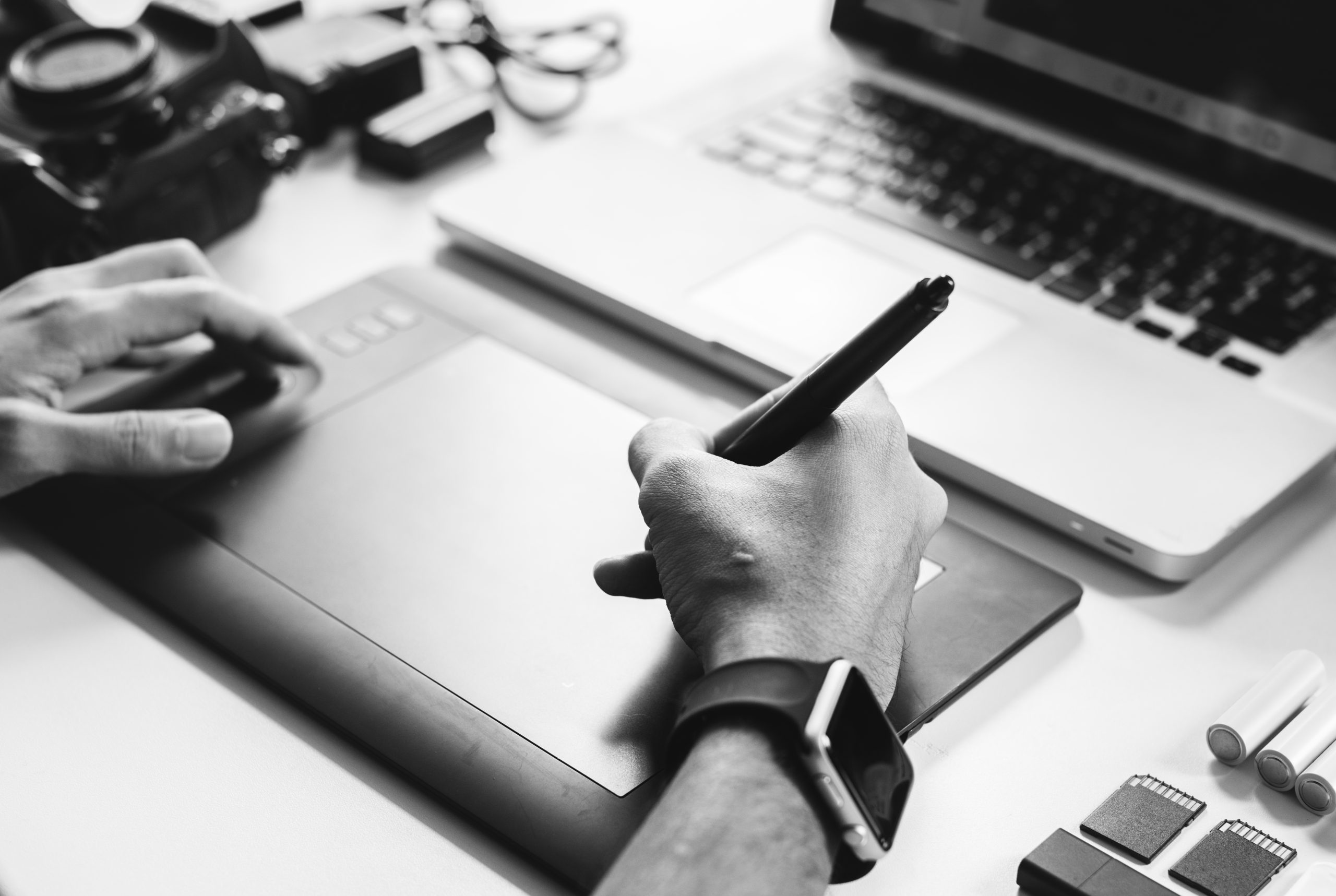 Designer utilisant une tablette graphique et un stylet devant un ordinateur portable, avec du matériel photo sur le bureau.