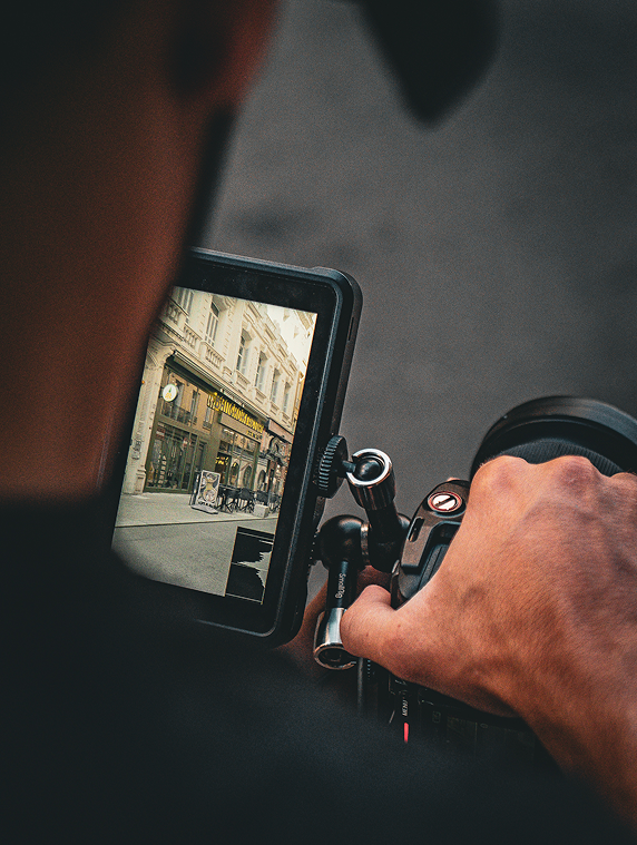 Vue rapprochée des mains d’un vidéaste tenant une caméra et regardant la scène filmée sur un écran externe montrant une rue commerçante.