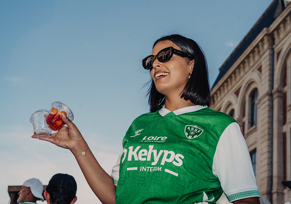 Femme souriante portant un maillot de football de l'ASSE et tenant un petit contenant de nourriture lors d’un événement en plein air.
