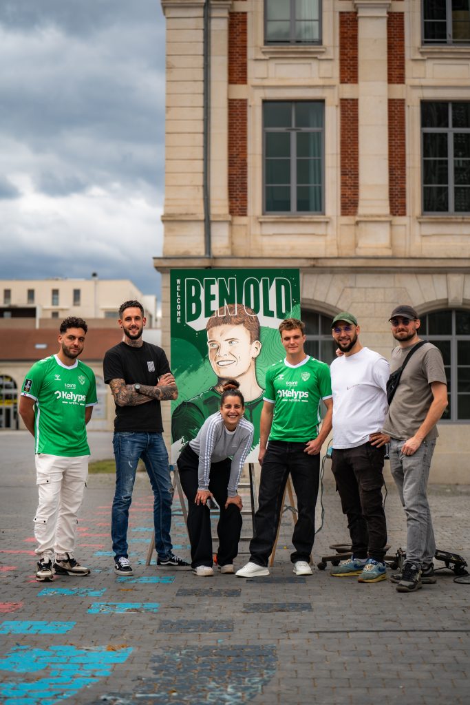 Sept personnes posant devant une grande affiche verte “Welcome Ben Old” sur une place, avec deux maillots de l’AS Saint-Étienne.