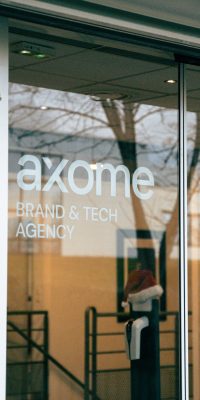 Façade vitrée d’un bâtiment avec les inscriptions « axome – Brand & Tech Agency » et « Welcome / Bienvenue » sur la porte.