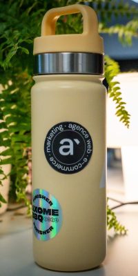 Gourde beige posée sur un bureau, avec un logo Axome et un autocollant « Axome BBQ 2024 », entourée de plantes et éclairée par une lampe.