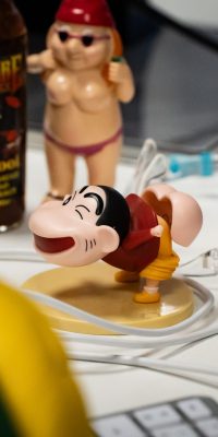Objets décalés sur un bureau : figurine cartoon au centre, deux figurines “gnomes” humoristiques derrière, une bouteille et une tasse, avec des câbles et adaptateurs.