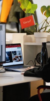Femme travaillant à son bureau dans un open space, avec un écran et un ordinateur portable ouverts, une gourde noire et une plante en pot.