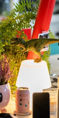 Bureau de travail avec lampe allumée, figurine de dinosaure, plantes et accessoires sur un poste informatique.