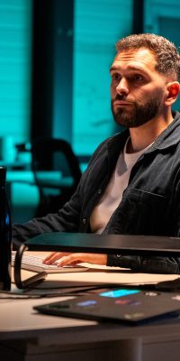Homme assis à un bureau, concentré devant un écran d’ordinateur, dans un open space éclairé en bleu/vert (plante, gourde et petite boule à facettes sur la table).