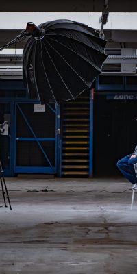 Plateau de tournage dans un entrepôt : un homme assis sur un tabouret est interviewé face à une caméra, avec un grand parapluie lumière.