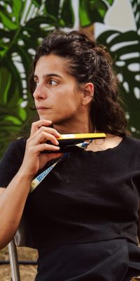 Femme assise en extérieur tenant un talkie-walkie, sur fond de décor végétal illustré.